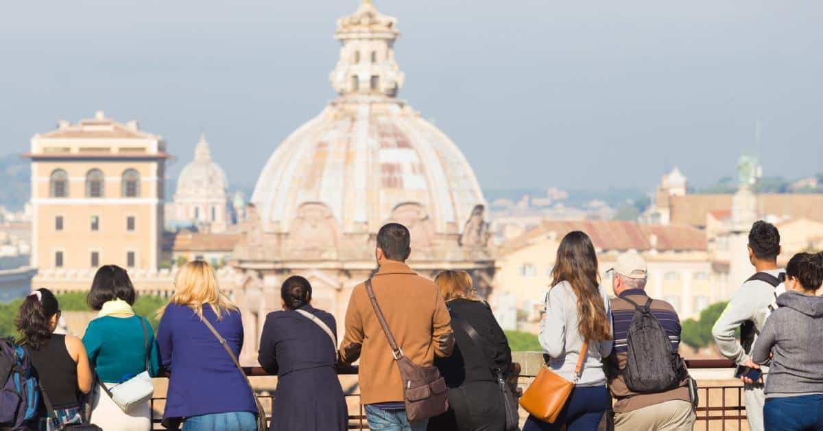 A group of tourists at the ledge of an overlook as they stare out at Rome, Italy from a distance. It's sunny outside.