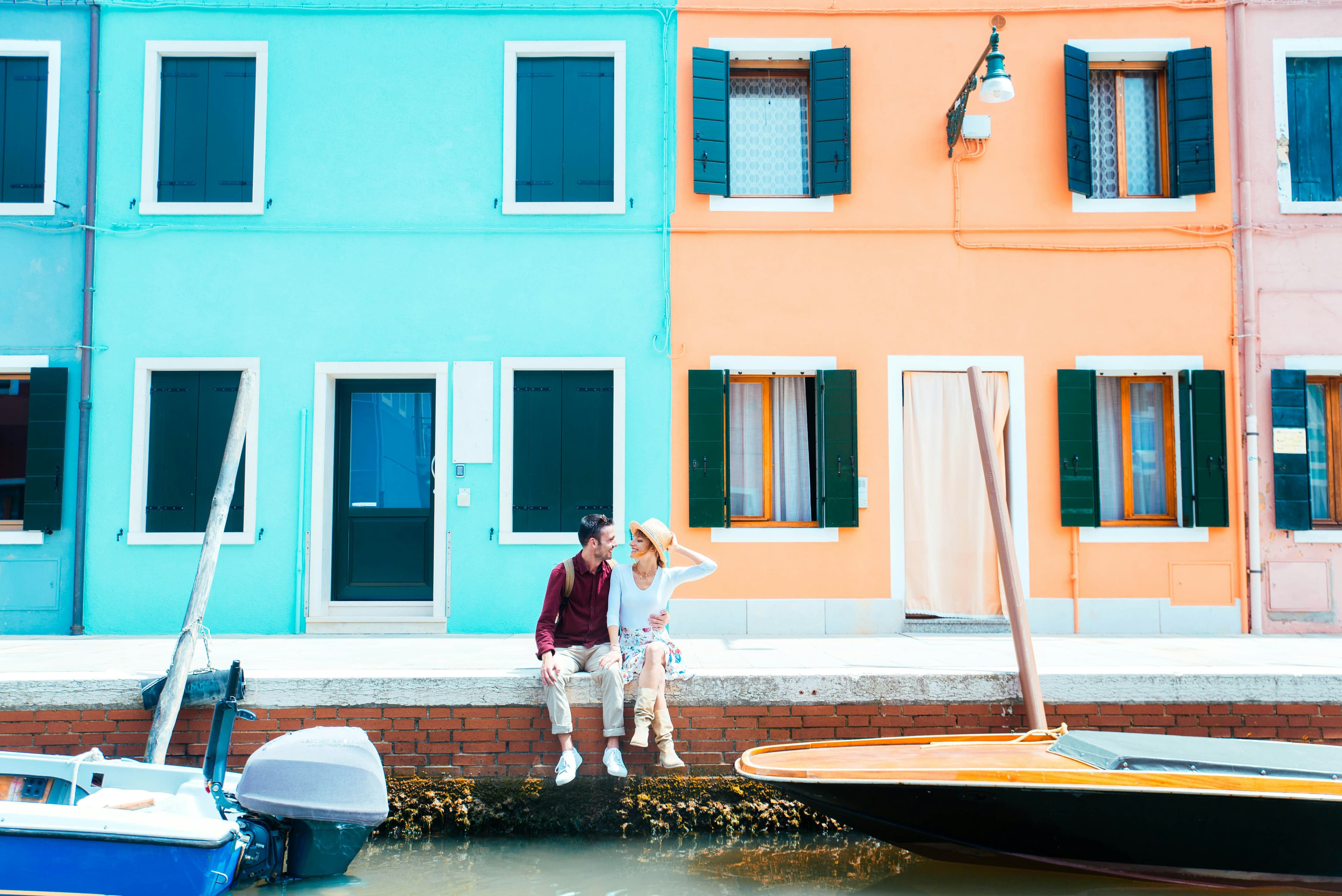 A couple sits together on a canal edge in front of bright blue and orange houses with green shutters—a sunny, cheerful scene that captures the charm of one of the Top Honeymoon Destinations 2024. Small boats float peacefully nearby.