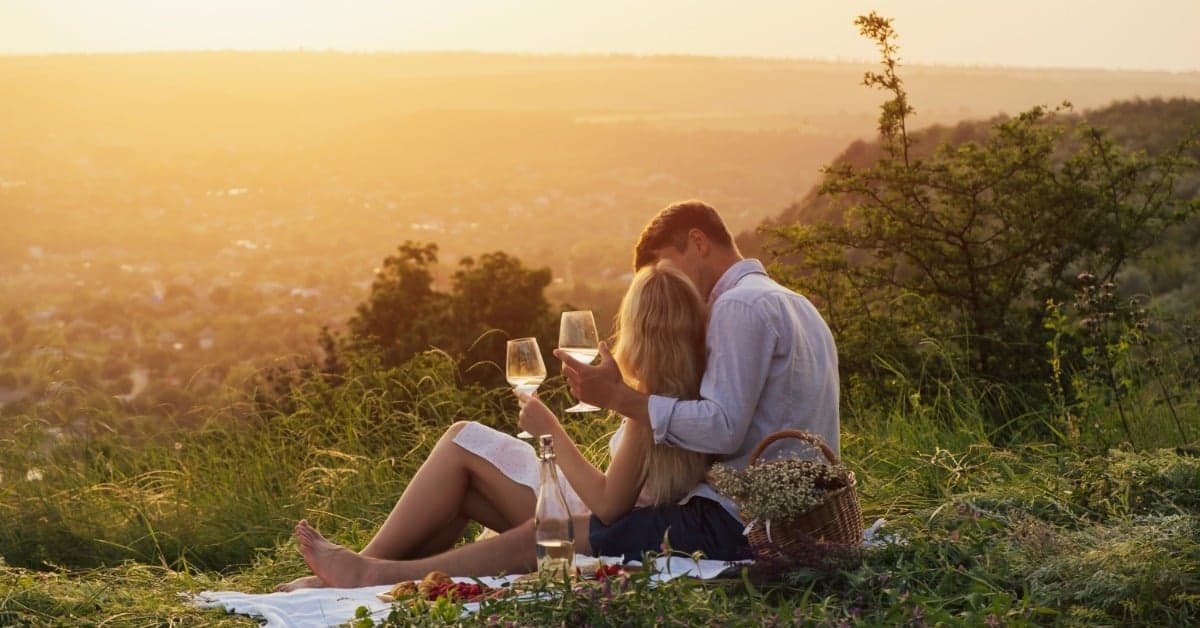 A couple sits on an Italian countryside hill as the sun goes down in the distance and they toast with wine glasses.