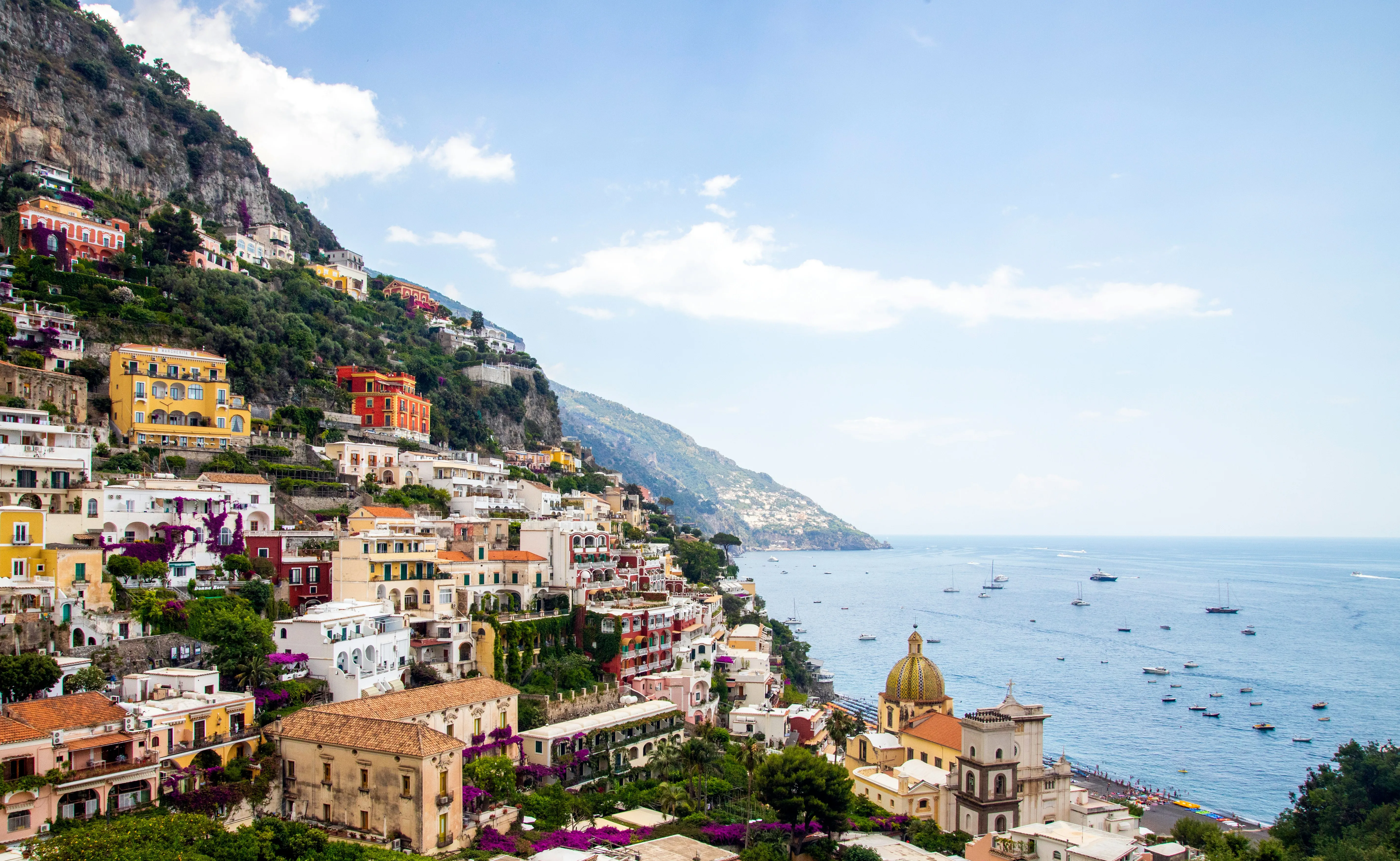 Colorful hillside buildings overlook the blue sea in Positano, Italy, with boats scattered in the water—echoing the dramatic cliffs found near Greece's Mount Olympus—lush greenery thrives under a bright, partly cloudy sky.
