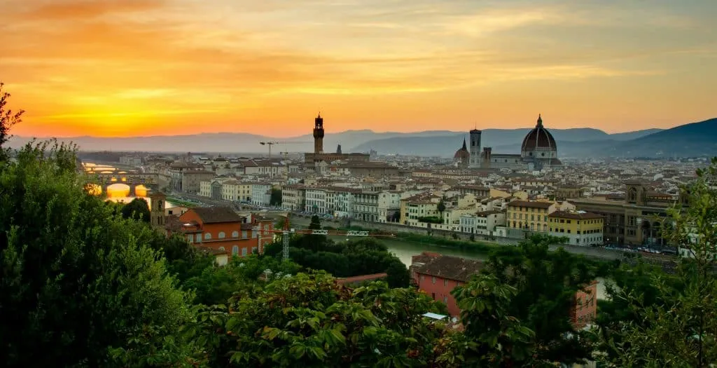 A scenic view of Florence at sunset, with the iconic Florence Cathedral and historic buildings highlighted. The Arno River gracefully flows through the city, as a glowing sky transitions from orange to blue, casting a warm light over the landscape—a true focus on Florence's timeless beauty.