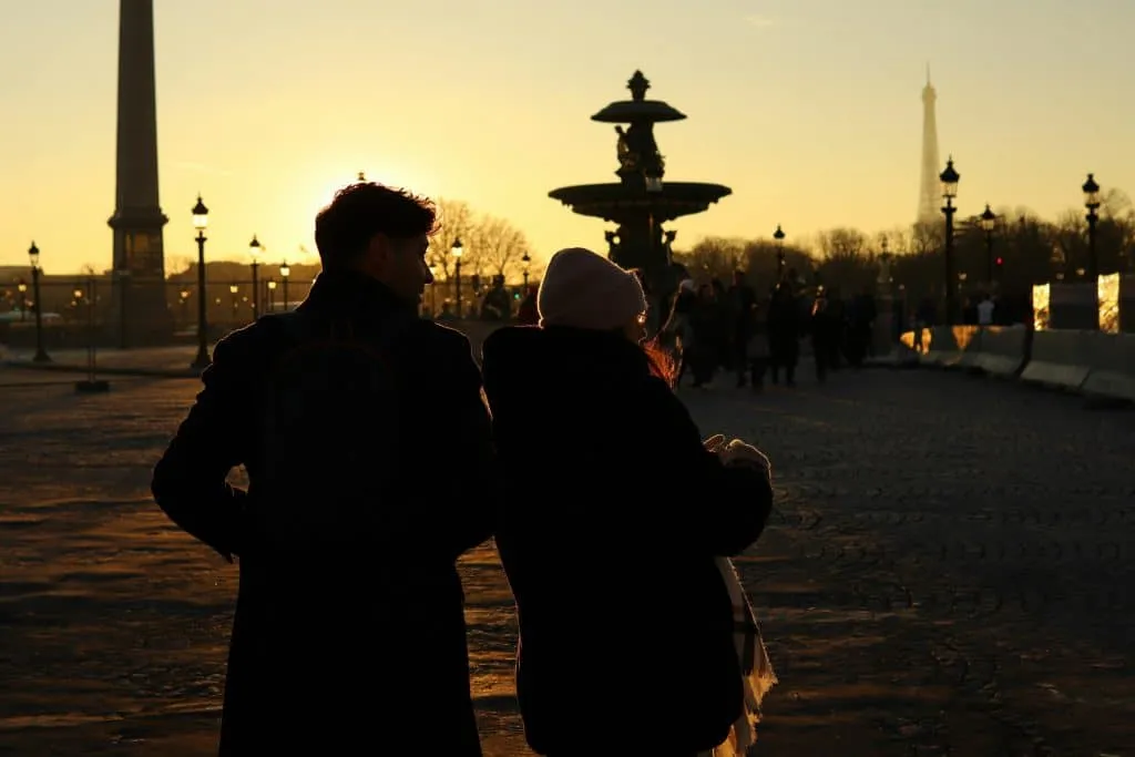 Two people standing in a public square at sunset, silhouetted against the sky. The Eiffel Tower is visible in the background, along with a large decorative fountain. The warm glow of the setting sun lights the scene. Place de la Concorde, Paris, France