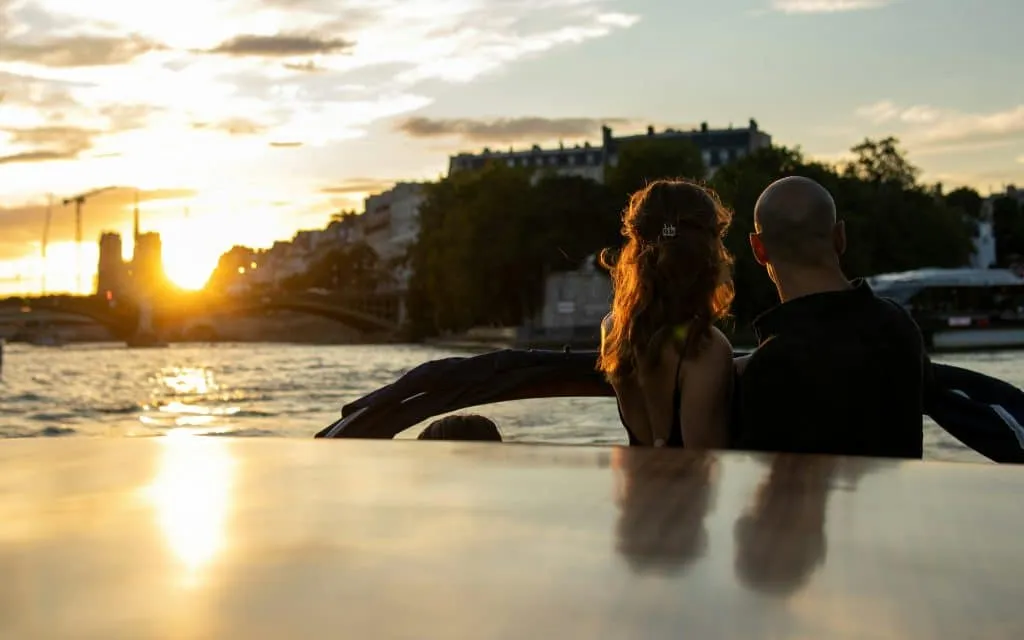 A couple sits at the back of a boat, watching the sunset over a river. The sun casts a warm glow on the water and silhouettes the couple. Buildings and trees line the riverbanks under a partly cloudy sky. Paris, France