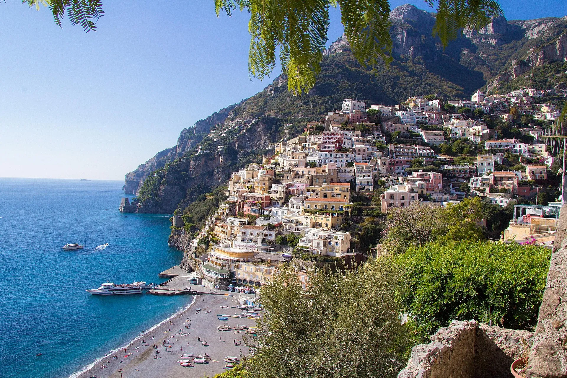 Colorful buildings cascade down a steep hillside toward a beach with umbrellas and boats on bright blue water in Positano, Italy, under a clear sky, with lush greenery—an iconic view perfect for Amalfi Coast custom itineraries.