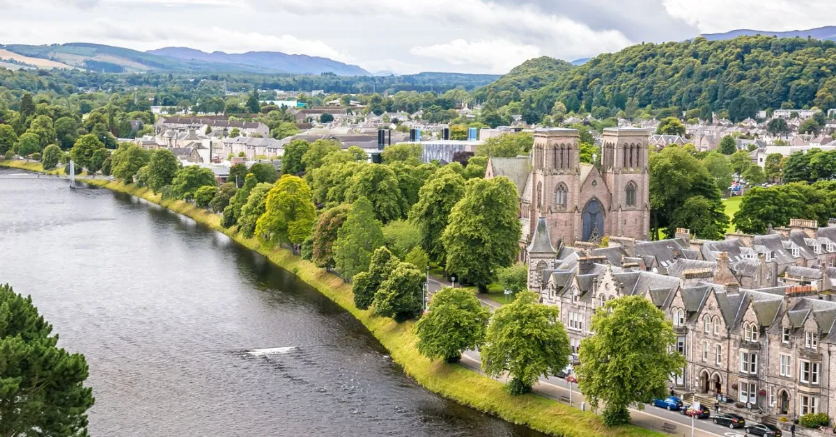 Inverness on the banks of the River Ness, with the red sandstone towers of St Andrew's Cathedral among trees and Highland hills beyond.