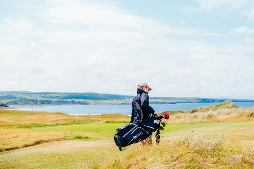 A person wearing a cap and jacket carries a golf bag across a grassy golf course, perfect for private group tours. The background showcases a scenic view of blue water and distant green hills under a cloudy sky.