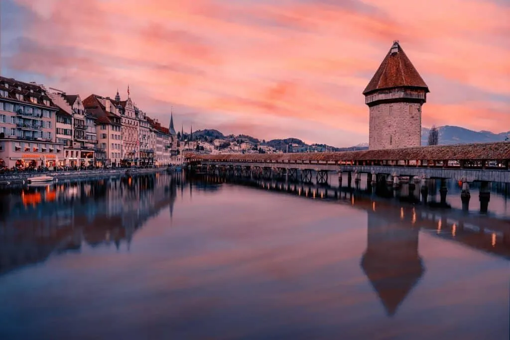 A serene view of a historic cityscape at dusk, featuring a stone tower with a conical roof by a river, reflecting the pink, orange, and blue sky. The water mirrors the vibrant sky and the charming buildings lining the riverbank.