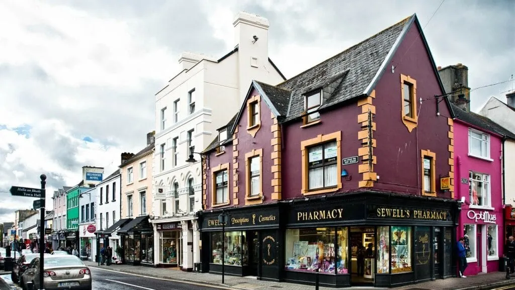 A street view in the quaint town of Killarney showcases colorful buildings, including a maroon and tan pharmacy on the corner. Cars are parked along the bustling street, while pedestrians stroll under a cloudy sky, adding charm to this picturesque scene.