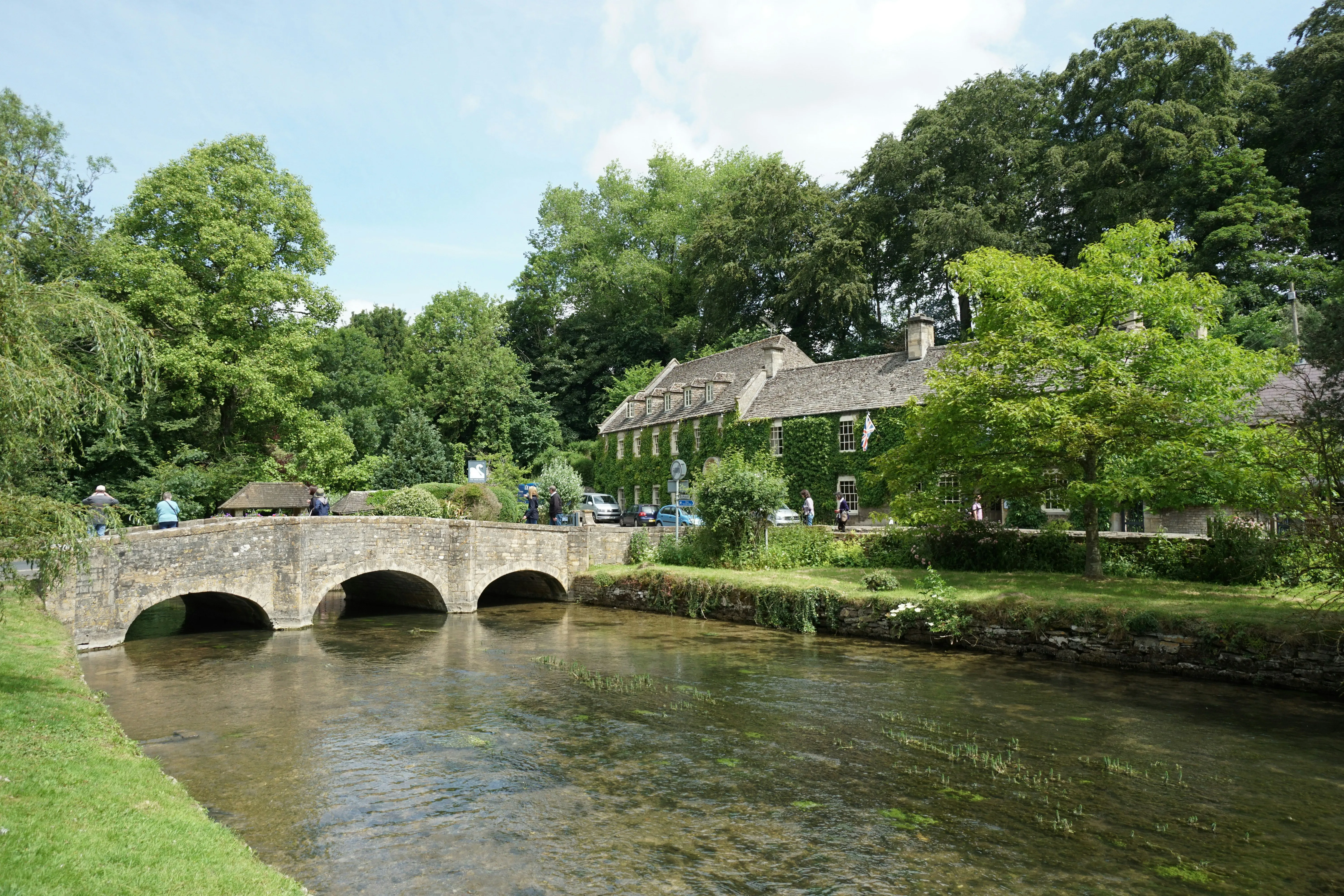 A stone bridge crosses a clear, shallow river in front of a row of old stone houses surrounded by lush green trees and grass on a sunny day. People are walking near the bridge and houses.