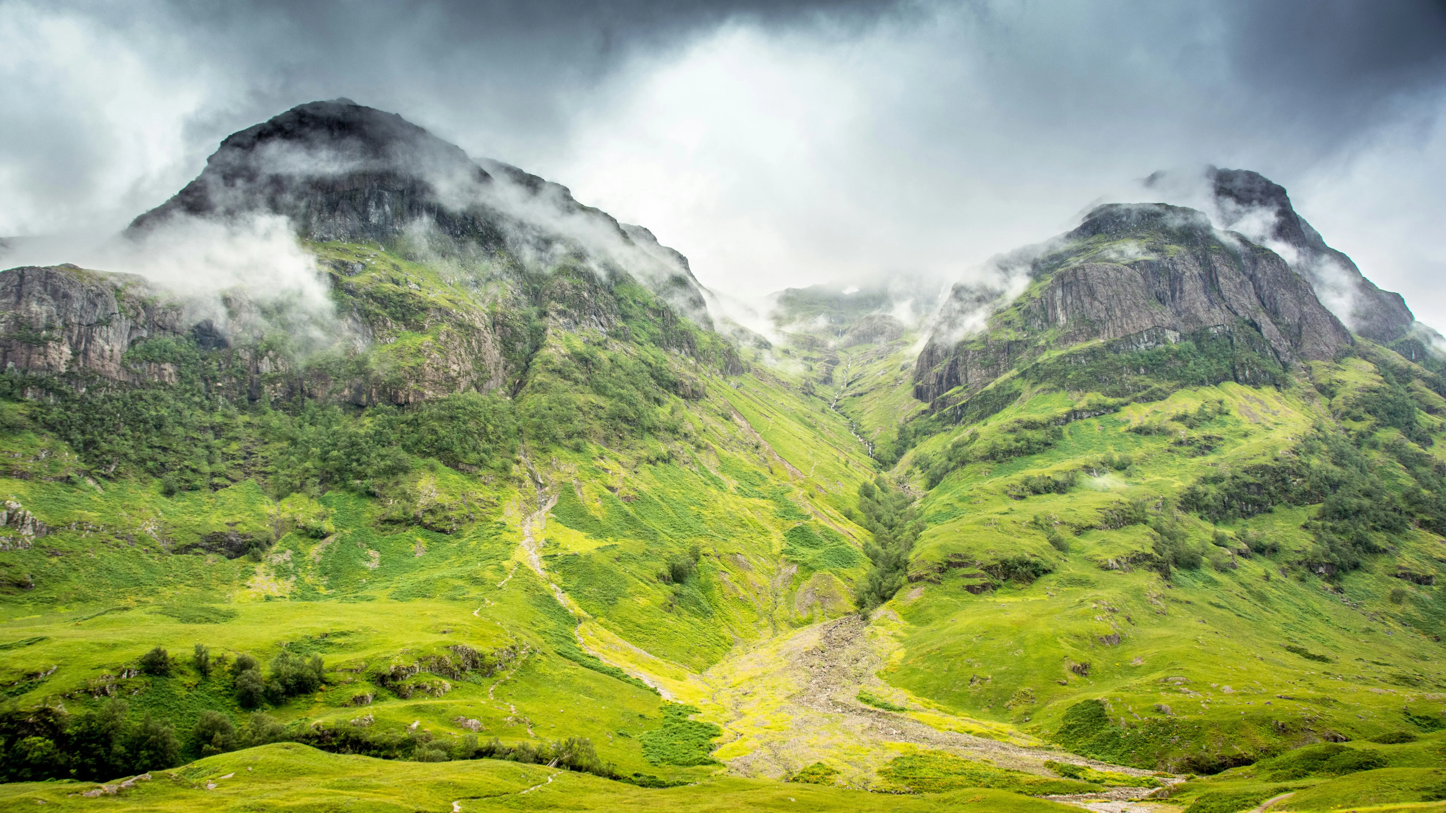 Mist and clouds drift over two steep, green mountains with rocky peaks and a lush valley below, under a dramatic, cloudy sky—evoking the landscapes seen where Game of Thrones was filmed.