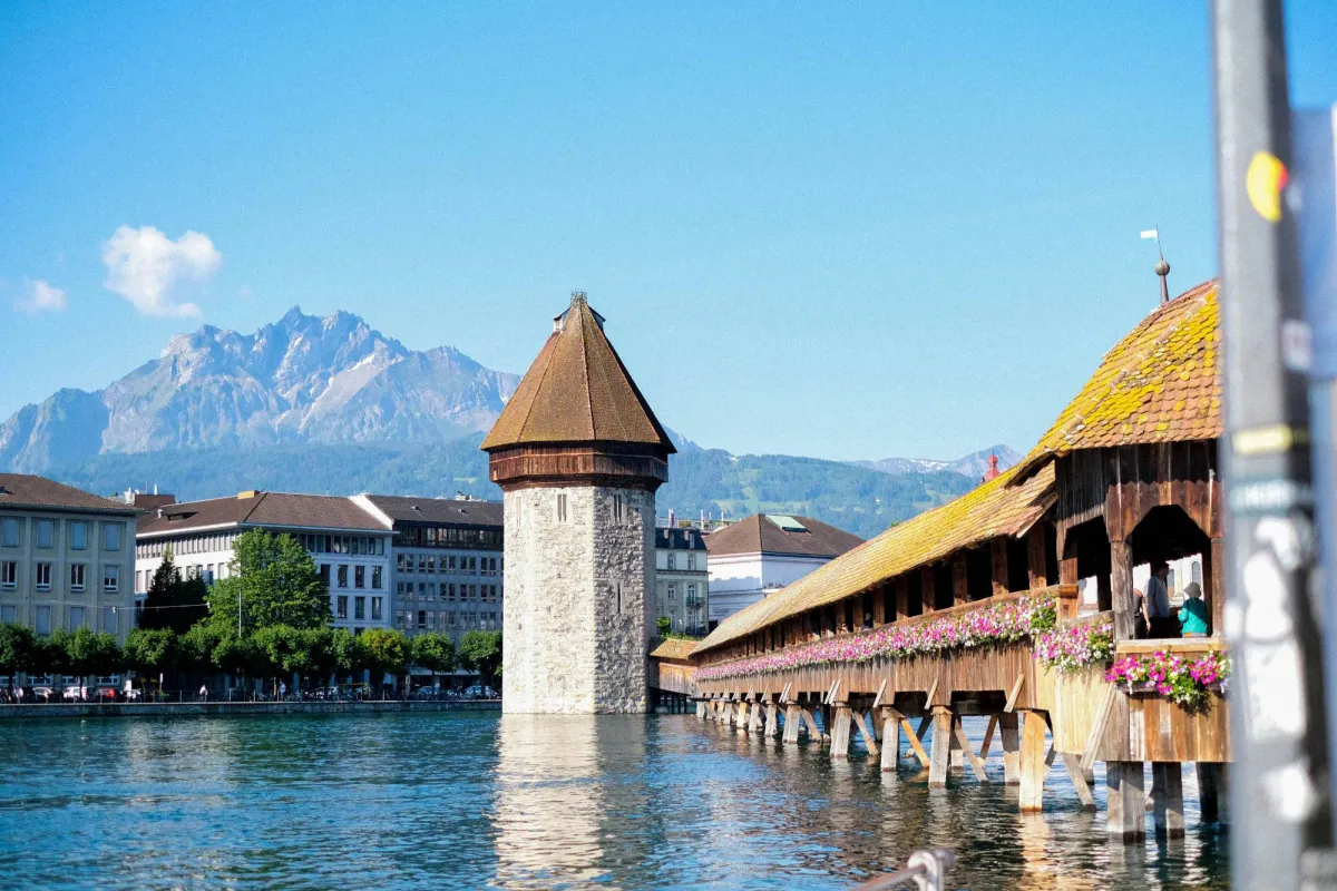Lucerne's wooden Chapel Bridge, hung with pink flower boxes, leads to the stone Water Tower on the Reuss River, with the jagged peaks of Mount Pilatus behind.