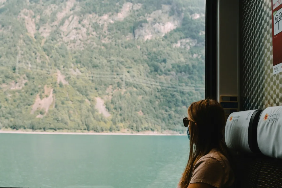 A traveler in sunglasses sits by the large window of a Bernina Express carriage, looking out at a forested mountain rising above a green Alpine lake.