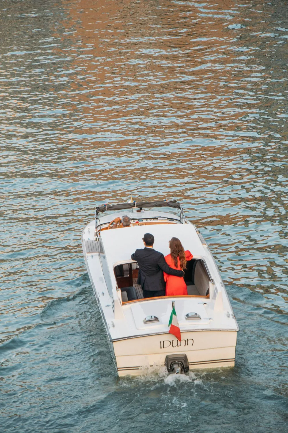 A couple standing arm in arm in a classic wooden motorboat on Lake Como, an Italian flag flying at the stern