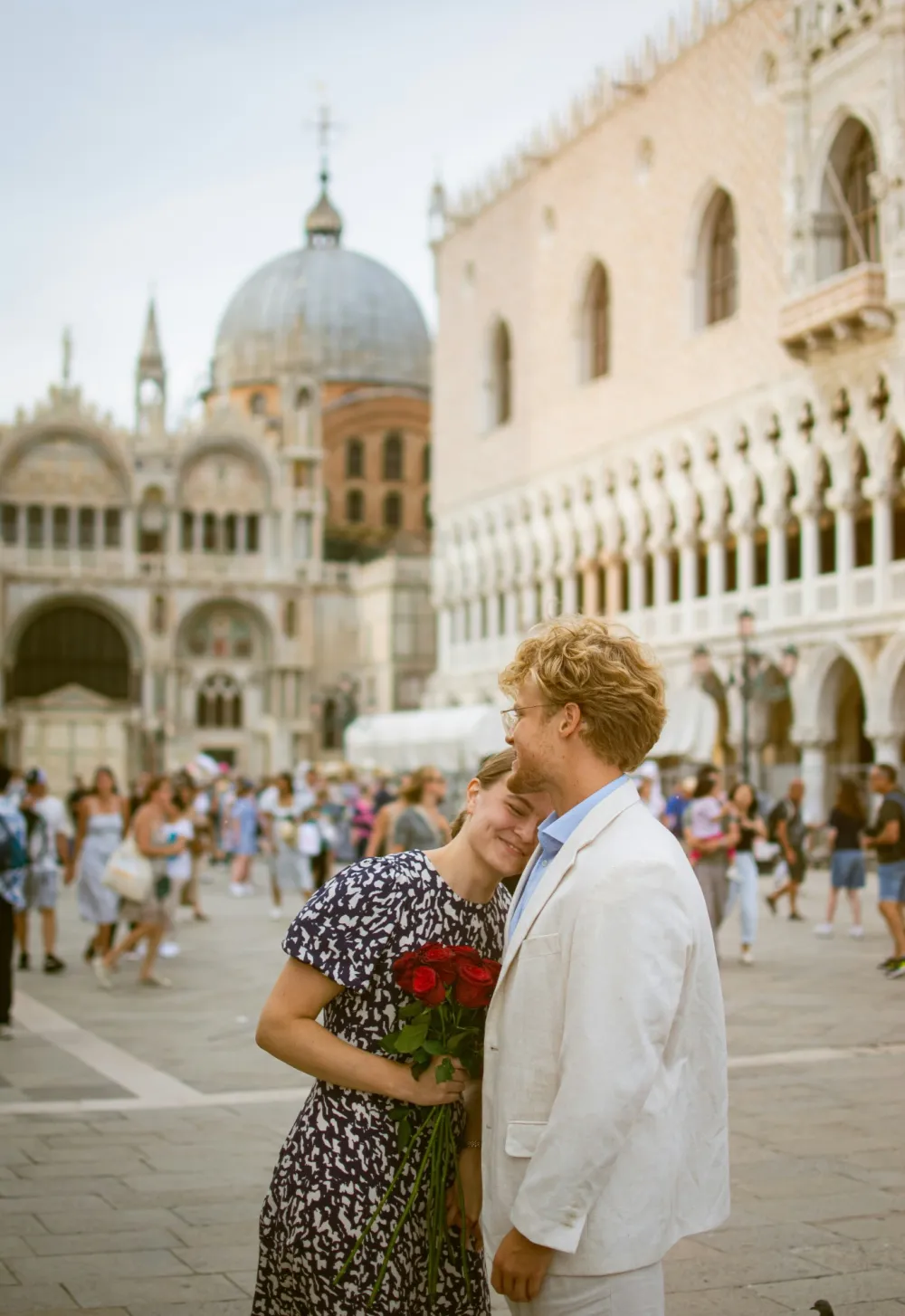 A smiling couple holding red roses in Venice’s Piazza San Marco, with St Mark’s Basilica and the Doge’s Palace behind them