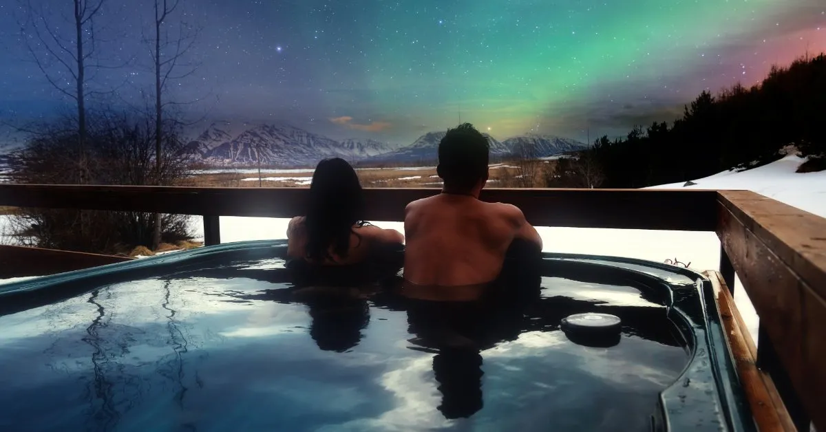 A couple relaxing in an outdoor hot tub on a snowy wooden deck, watching green northern lights over snow-capped mountains in Iceland.
