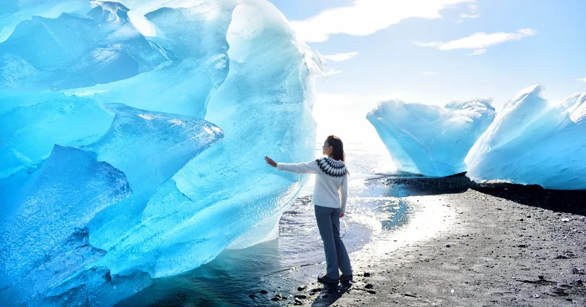 A traveler in an Icelandic wool sweater reaches toward a towering block of blue glacier ice on the black-sand shore near Jökulsárlón.