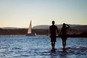 Couple standing in the water holding hands with a sailboat in background