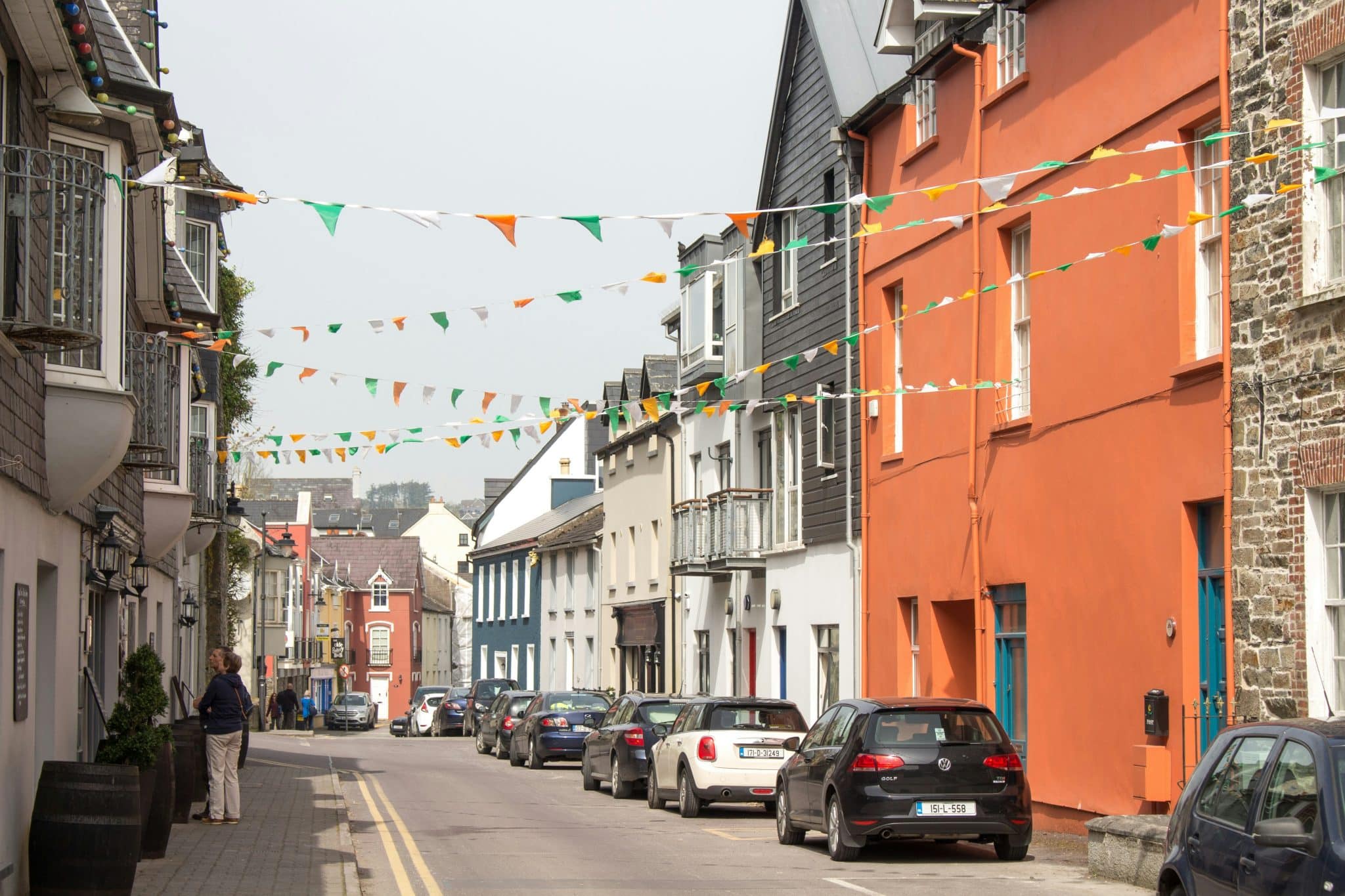 A street lined with colorful buildings, cars parked along the side, and strings of triangular bunting overhead captures the charm of elope worthy Ireland. A few people stroll on the sidewalk beneath an overcast sky.