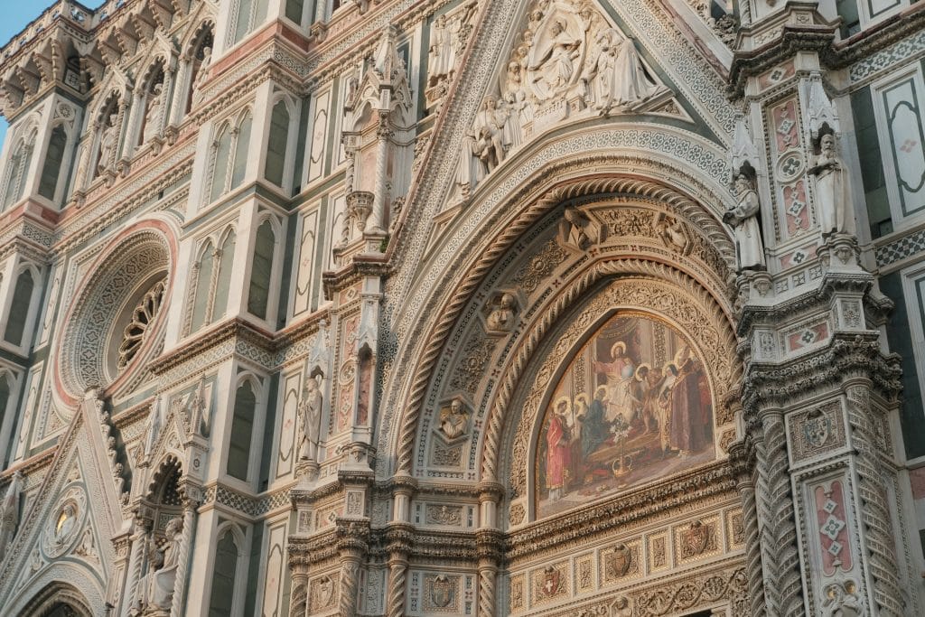 Close up of the Florence cathedral facade reminiscent of the architectural grandeur found in Rome, featuring elaborate carvings, arches, and detailed artwork. The intricate design includes religious figures and ornamental patterns in muted tones.