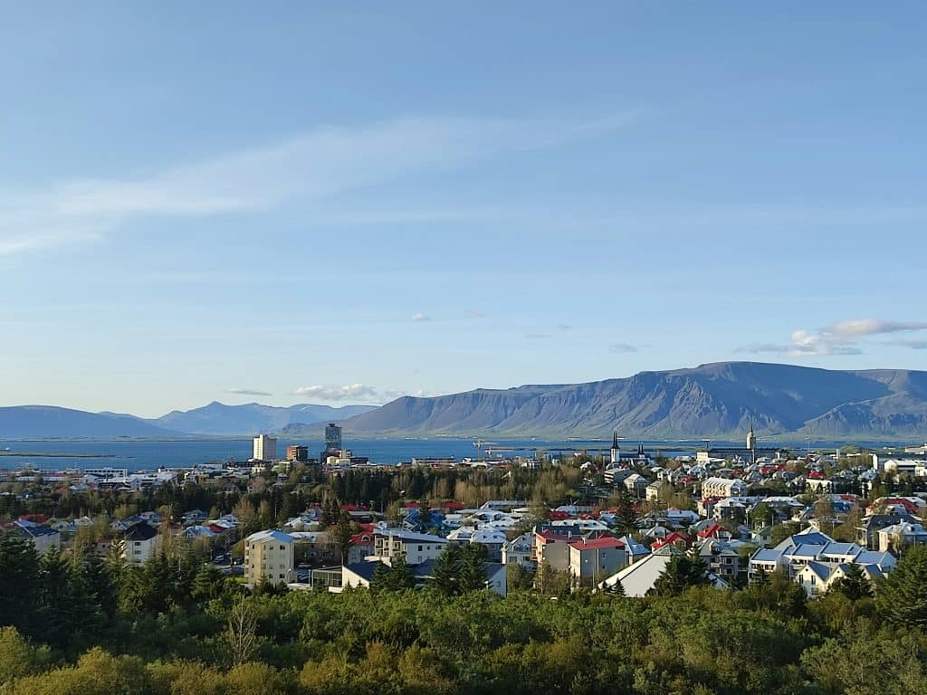 A scenic view of Reykjavík, a coastal city where colorful rooftops and green trees mingle. In the background, the calm waters meet the rugged mountains of the North under a clear blue sky.