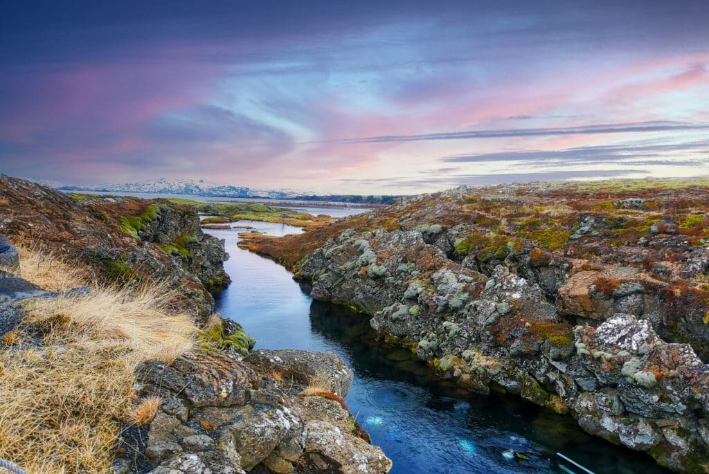 Scenic view of a narrow, clear stream flowing through rocky terrain with sparse vegetation. The sky is painted with soft hues of pink, purple, and blue as the sun sets, reflecting on the water. Distant snowy mountains are visible on the horizon. Sulfra Fissure, Thingvellir, Iceland