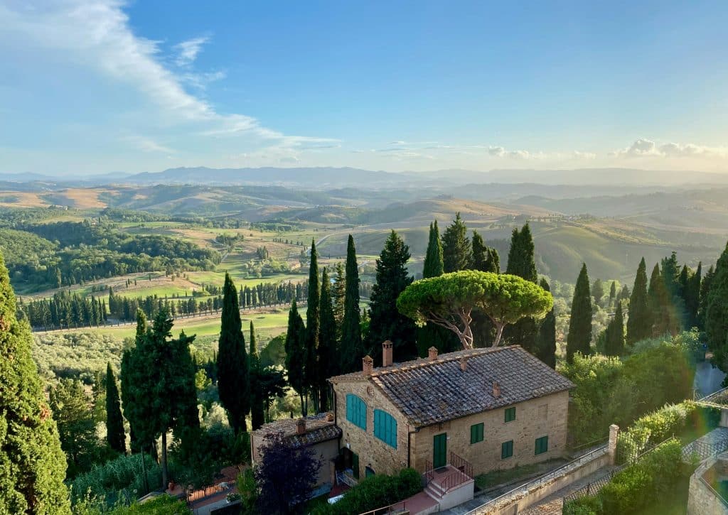 Scenic countryside view featuring a stone house with a tiled roof amid tall cypress trees, overlooking rolling green hills under a clear blue sky. Experience the best of Italy as clouds gently dot the horizon.