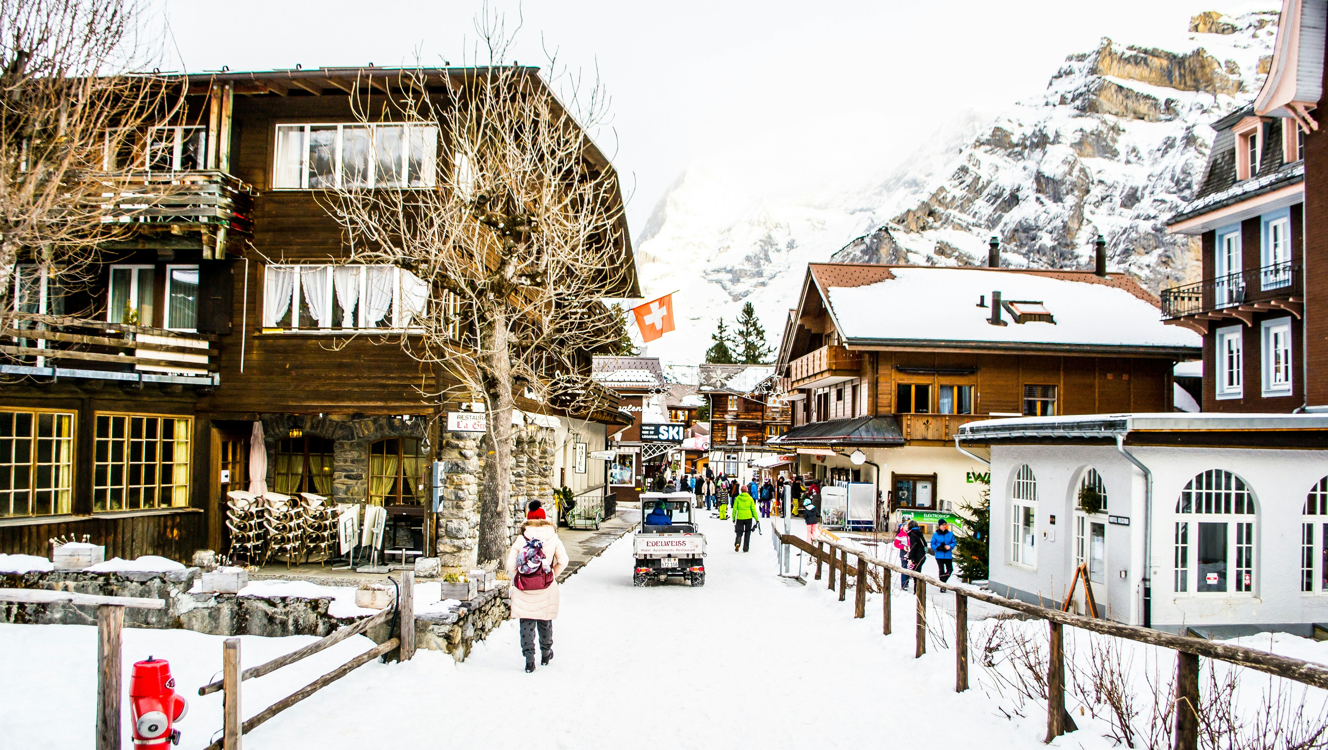 A snowy street in a Swiss mountain village with wooden chalets, a Swiss flag, and people walking beneath snow covered rooftops; mountains rise in the background—an ideal setting for those exploring Winter Olympics 2026 Travel Packages.