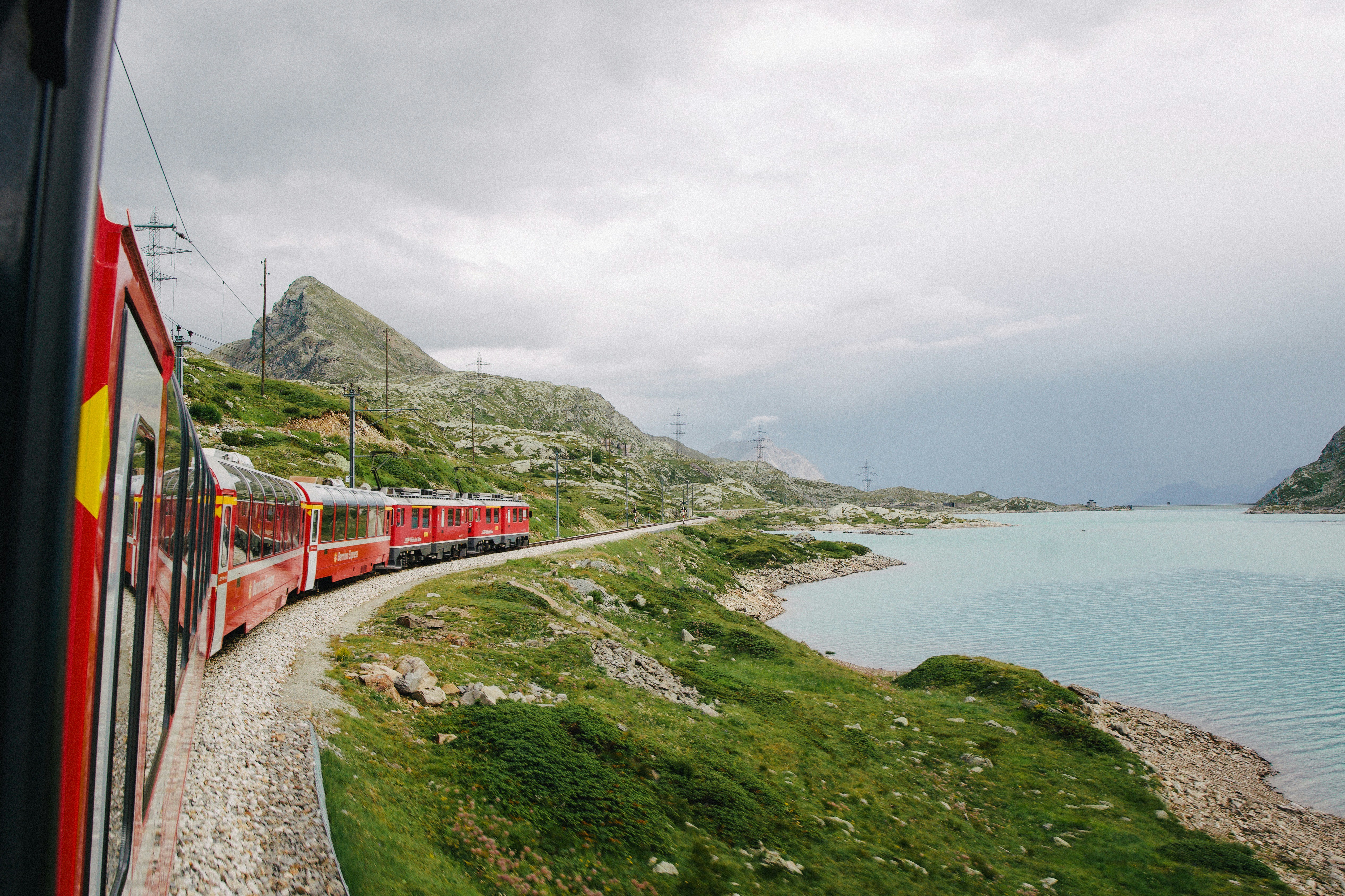 A red Glacier Express train curves along tracks by a turquoise lake in Switzerland, surrounded by green hills and rocky mountains under a cloudy sky.