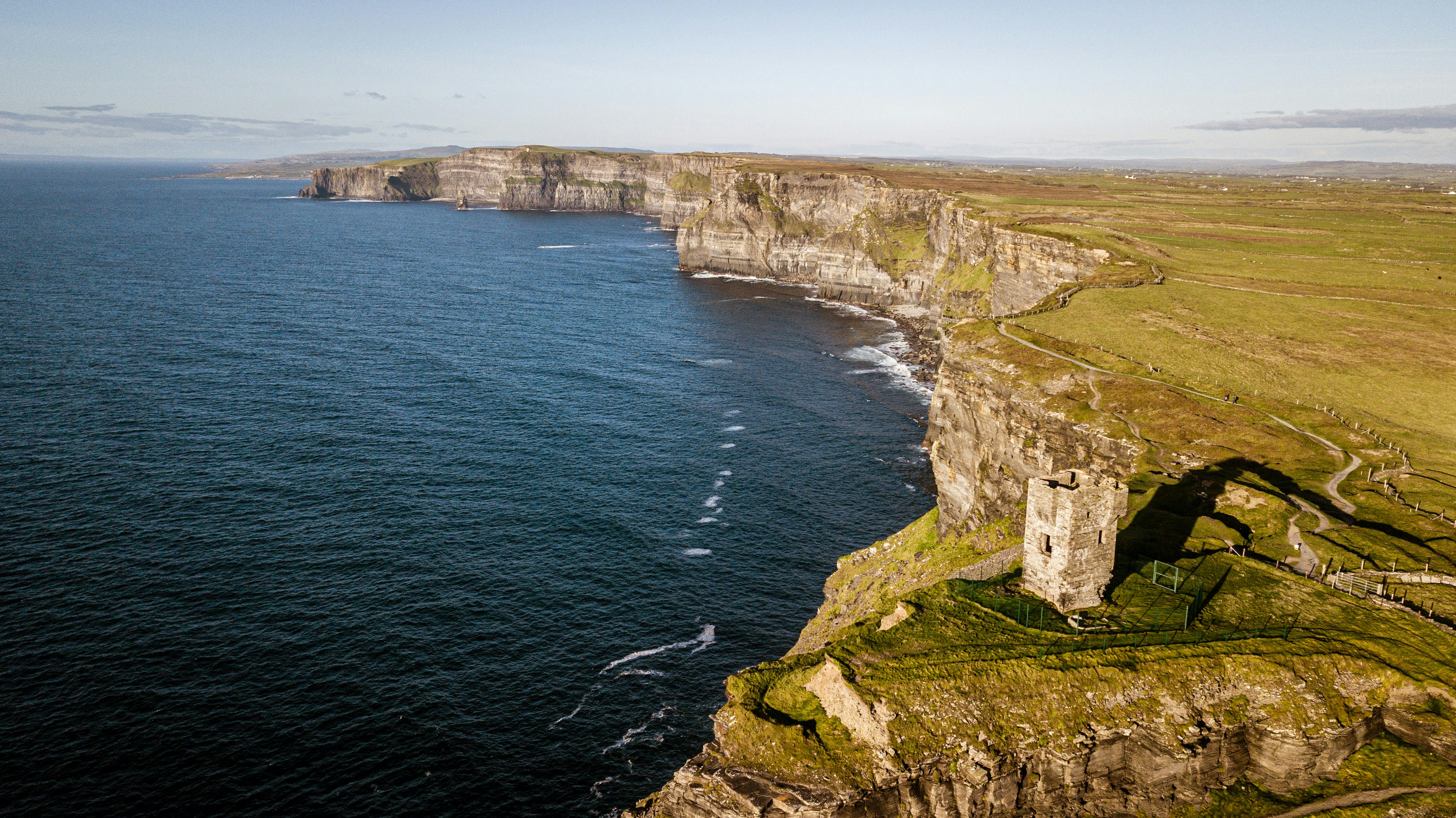 Aerial view of rugged sea cliffs with a stone tower on the edge, green grass, and the ocean stretching into the distance under a clear sky—one of the Best Places to Visit in Italy for 2025.
