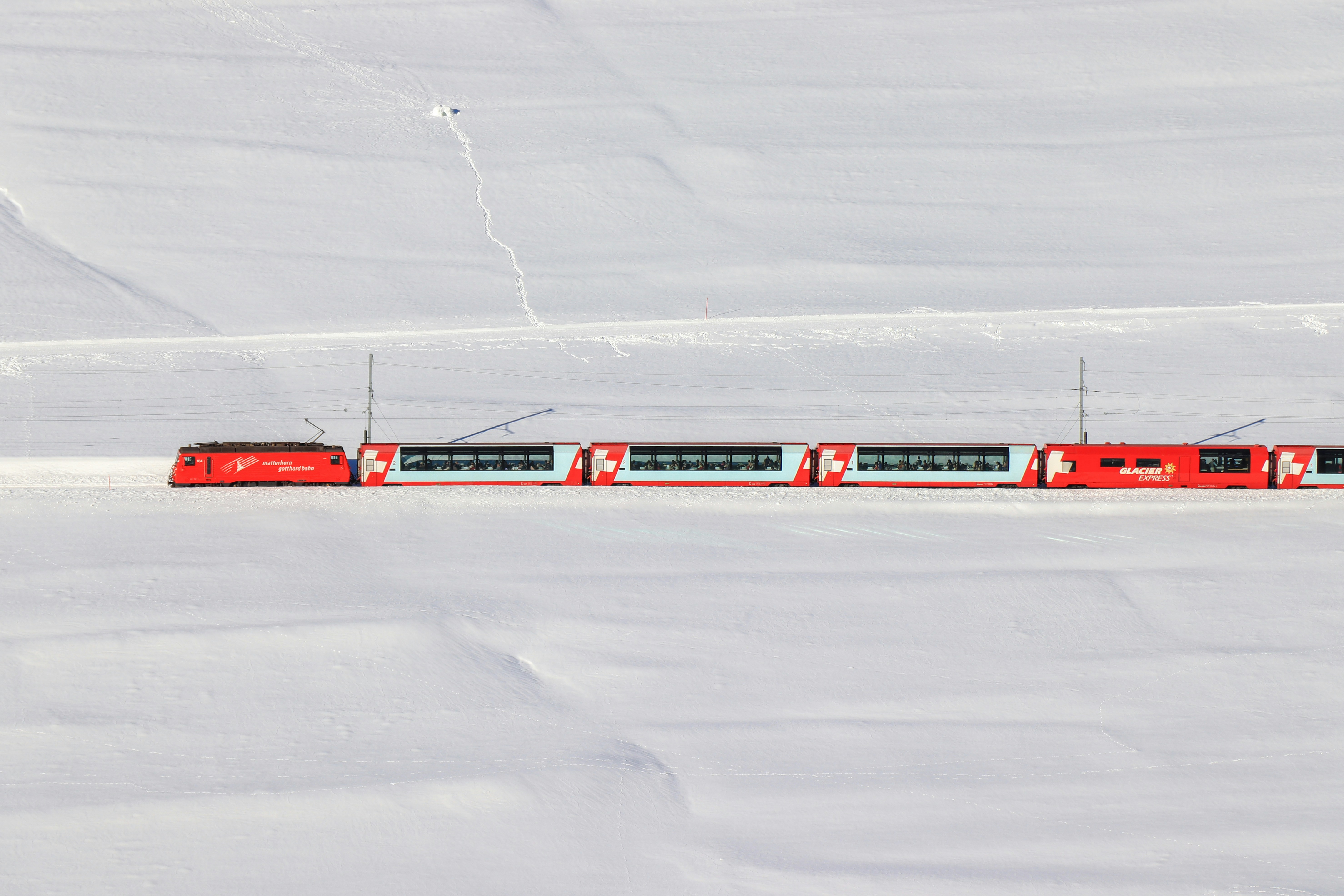 A bright red passenger train travels through a snowy, white landscape in Iceland, creating a striking contrast against the snow covered ground—much like the country’s dramatic scenery shaped by its many volcanoes. The scene is viewed from above, with the train as the focal point.