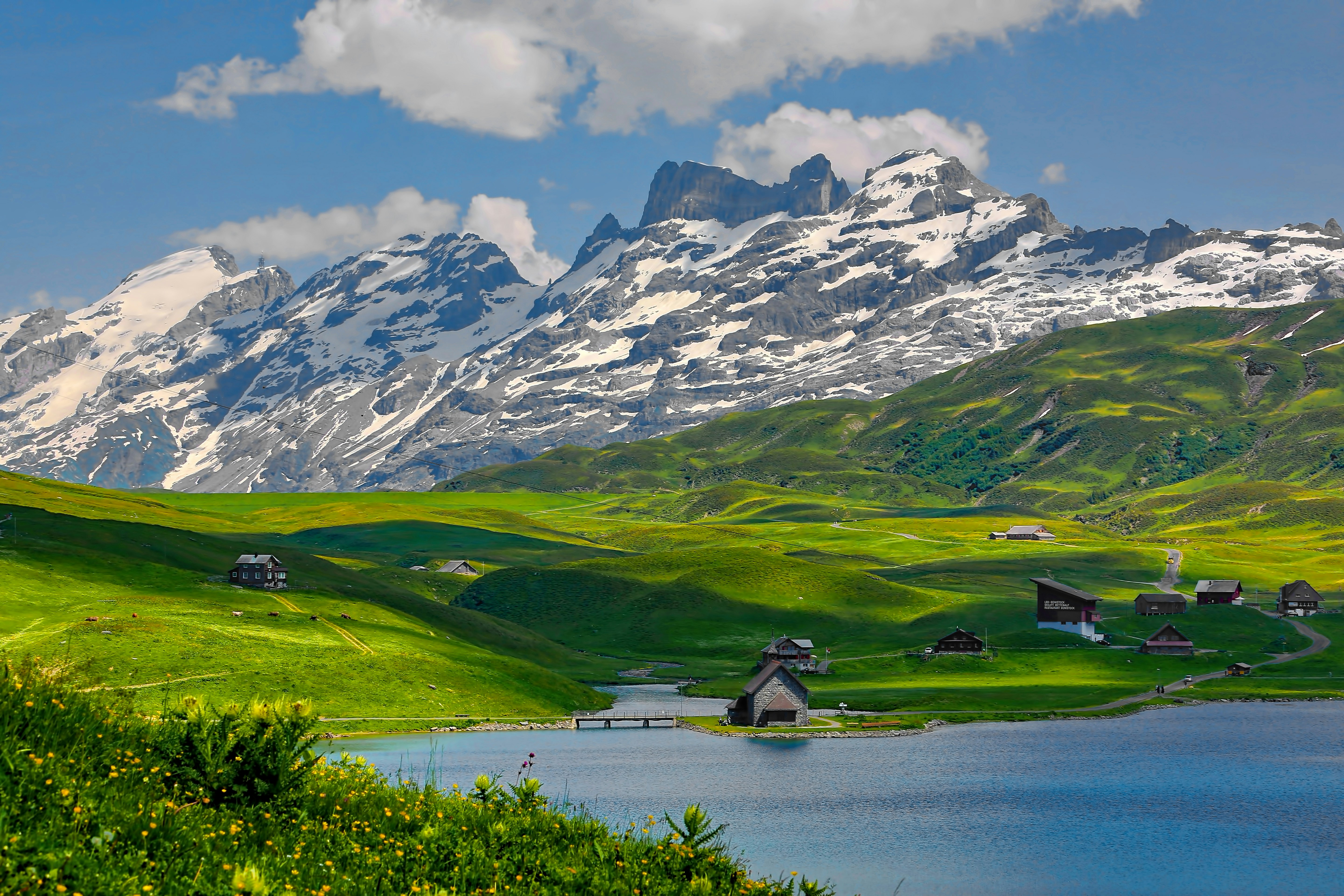 A scenic landscape featuring green hills, a blue lake, scattered houses, and snow capped mountains under a partly cloudy sky.