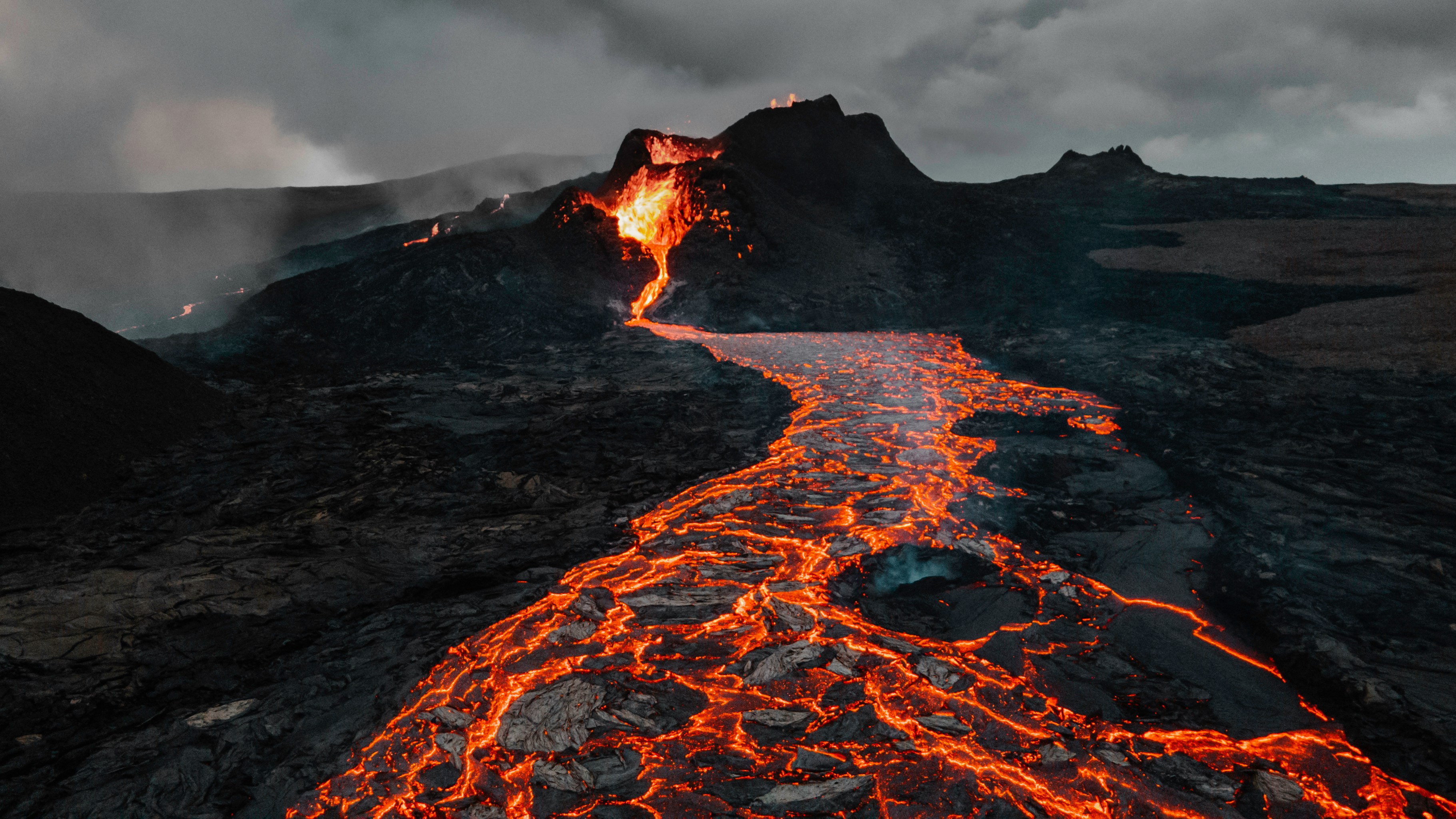 A glowing river of molten lava flows from the crater of an erupting volcano, cutting through dark, rocky terrain under a cloudy, smoky sky.