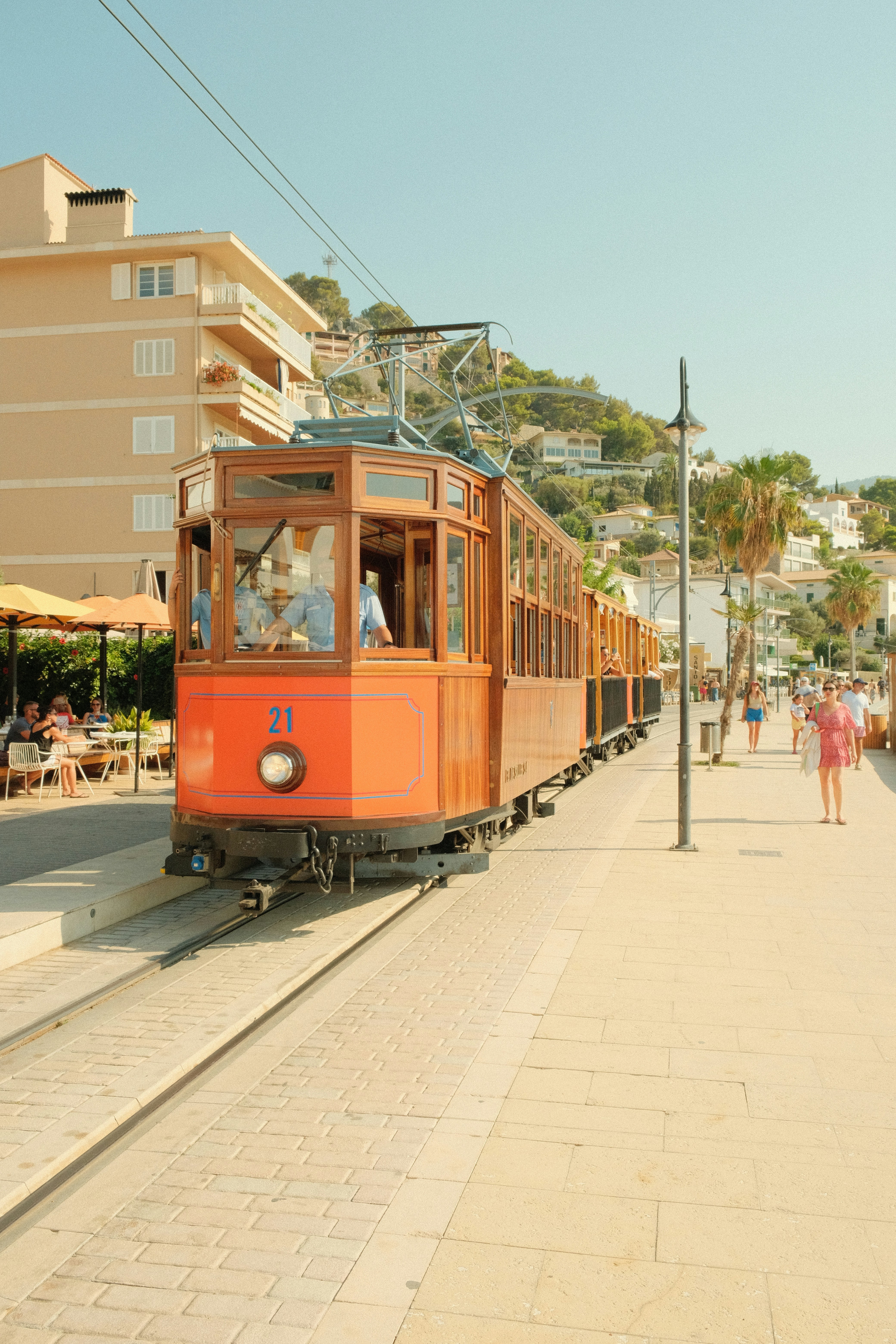 A vintage wooden tram moves along tracks beside a sunny promenade, with people walking and sitting at outdoor café tables—reminiscent of relaxing after visiting castles in Scotland to tour for free. Mediterranean style buildings and palm trees complete the scene.