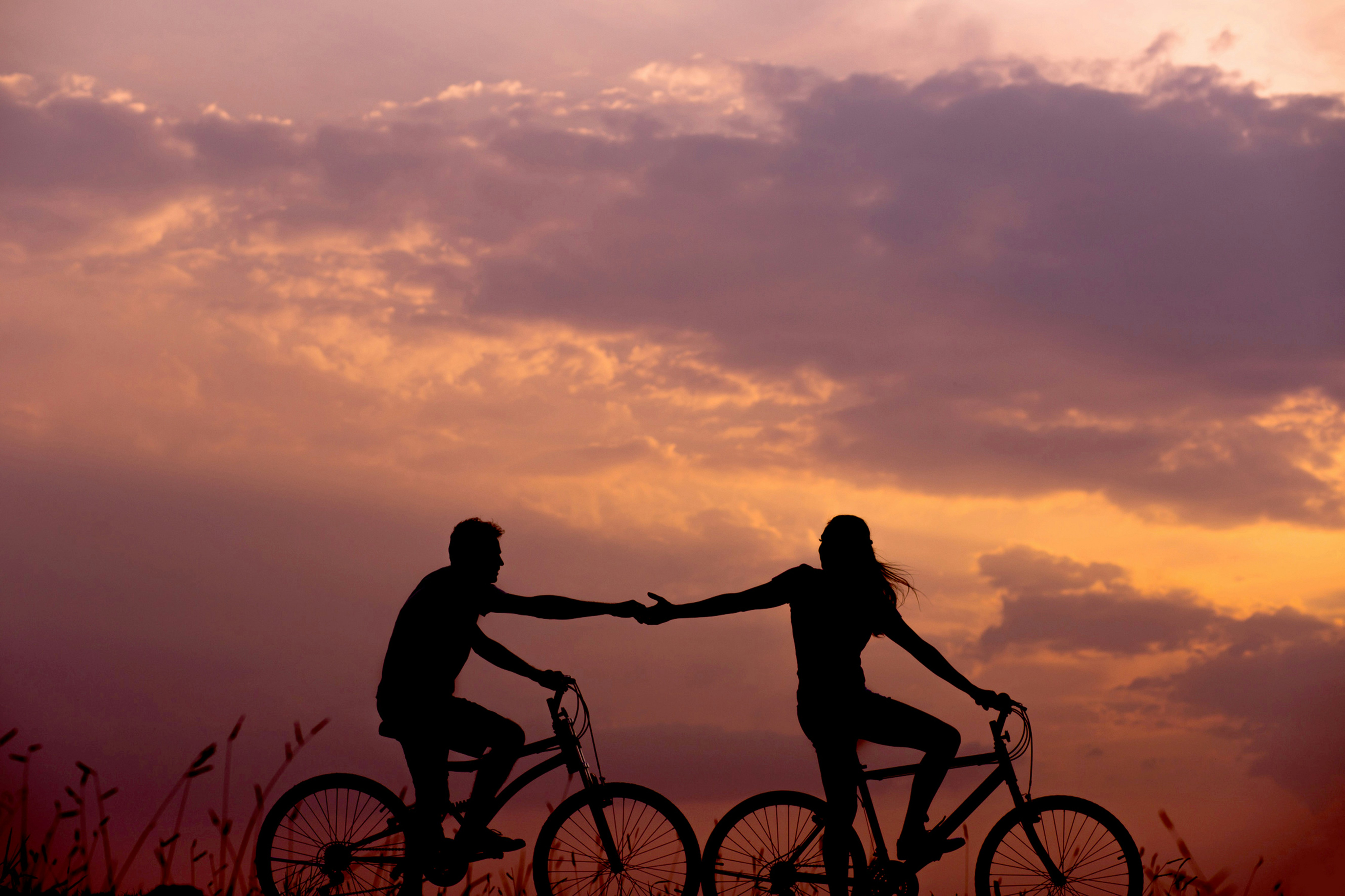 Two people on bicycles reach out to hold hands while riding at sunset, silhouetted against a colorful sky—capturing the spirit of adventure and togetherness found when learning how to plan a multi country tour in Europe.