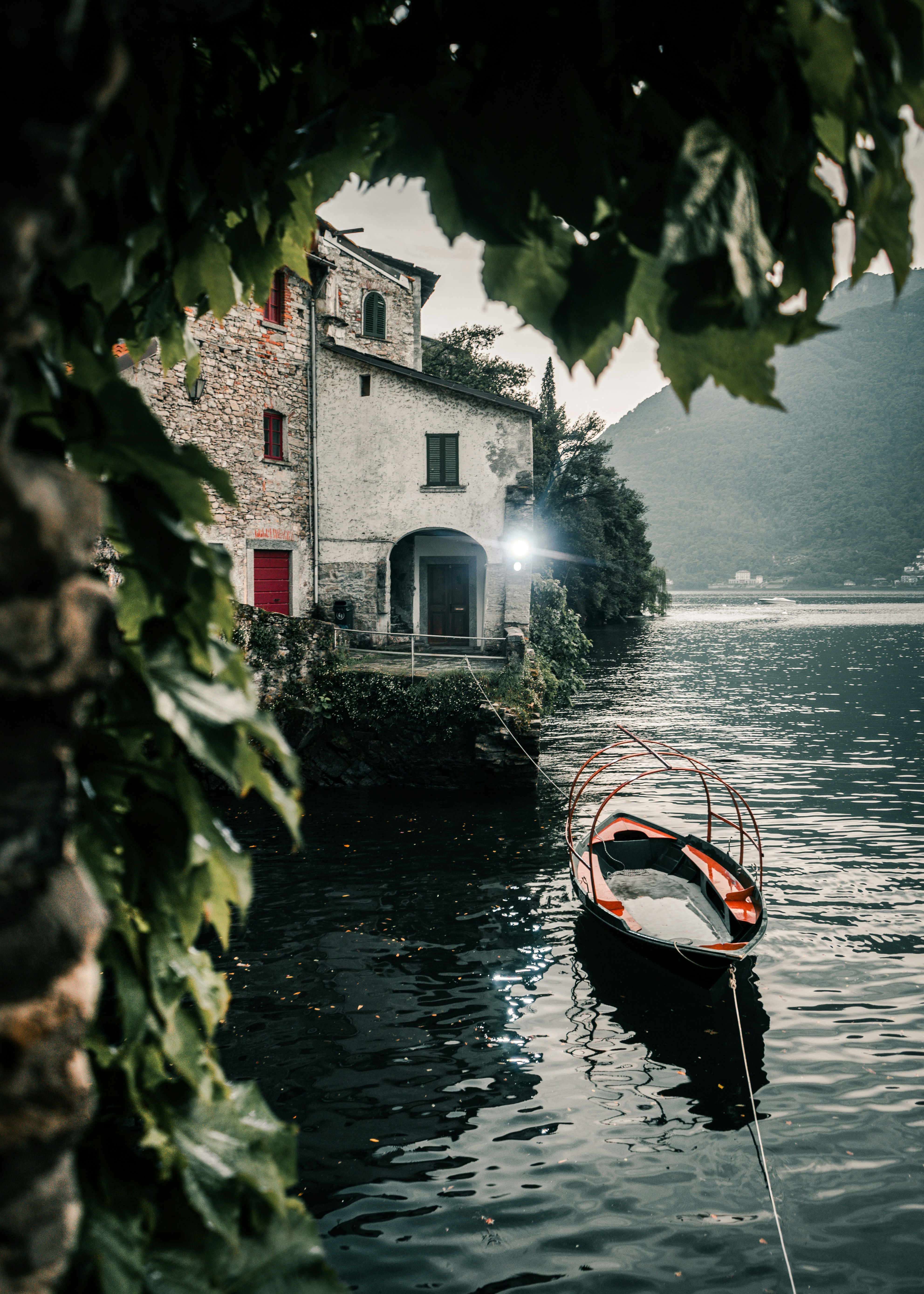 A small wooden boat is tied to the shore beside an old stone house on a calm lake, surrounded by lush greenery and misty mountains—evoking the spirit of adventure found in Vikings in Dublin 2025 Travel Packages. Leaves frame the serene scene.