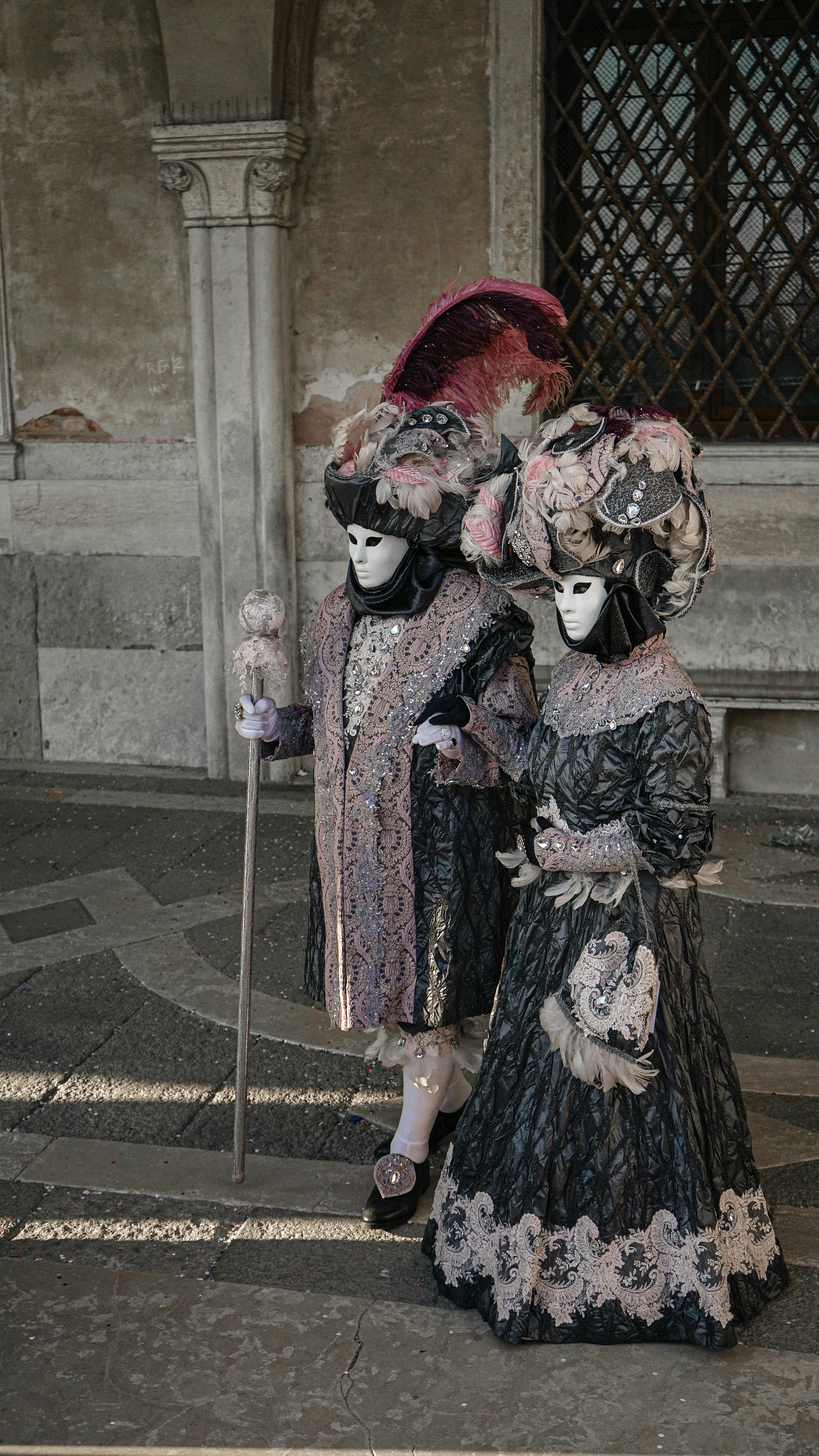 Two people in elaborate, ornate costumes and decorative masks stand side by side at the Venice Carnival. They wear richly detailed garments with lace and feathers, one holding a staff, against an old stone wall and barred window.