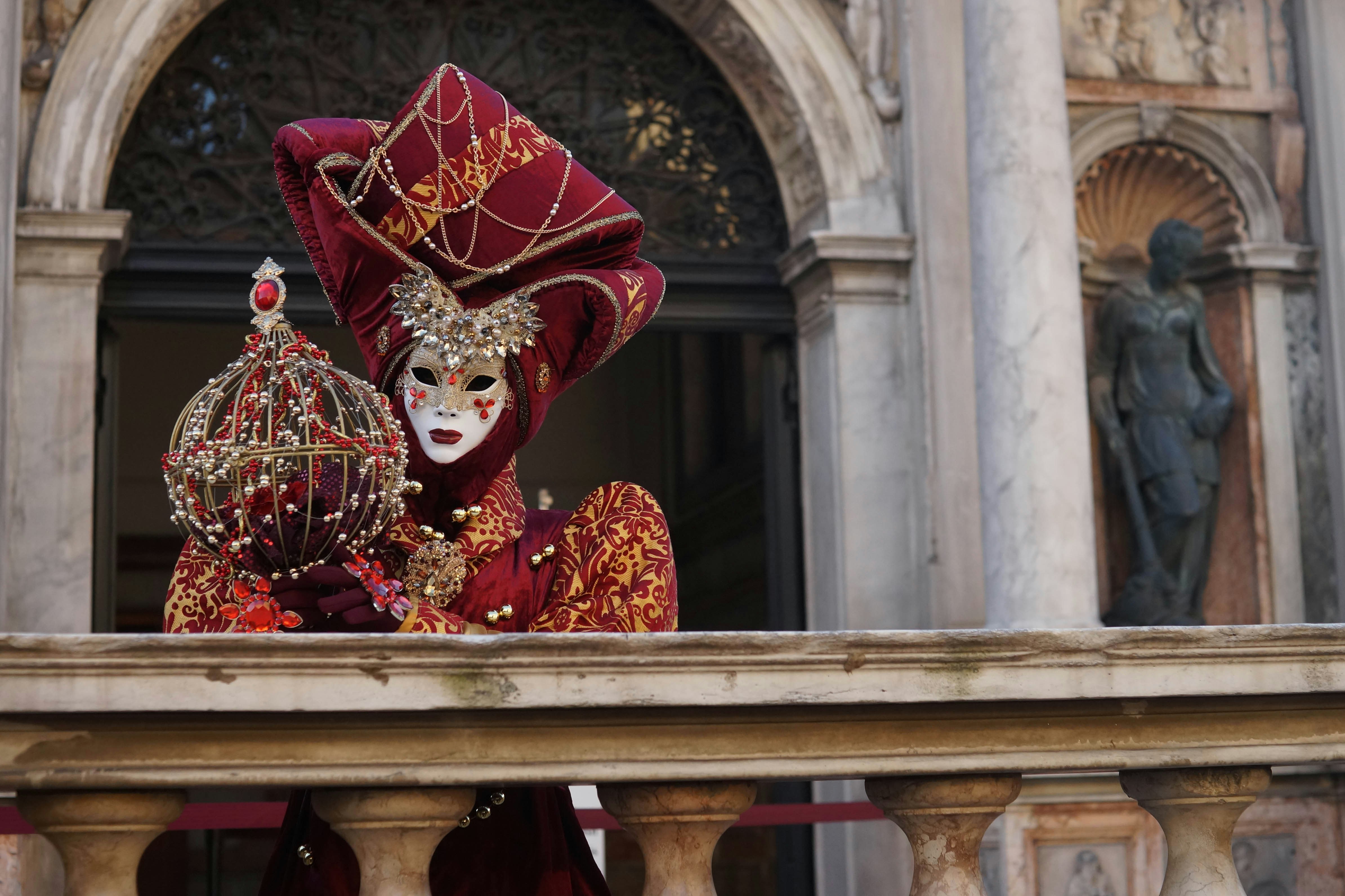 A person in an ornate, red Venetian carnival costume and mask stands behind a stone railing, holding an elaborate spherical ornament—bringing a touch of gaff, a quintessential Irish slang for 'home,' to the historic building and statue in the background.