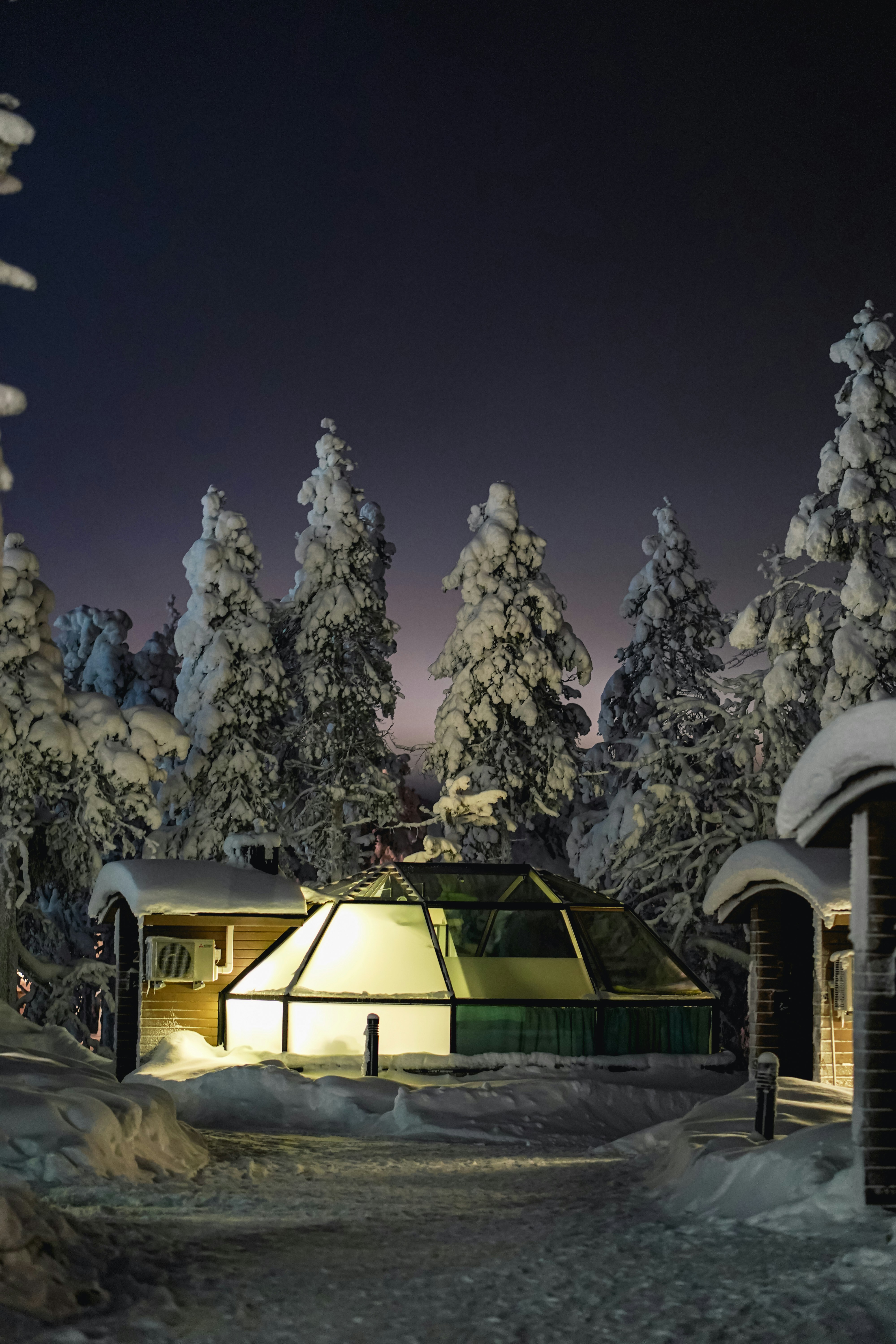 A glass igloo glows with warm light amid snow covered trees at night, creating a cozy atmosphere reminiscent of the Best Honeymoon Locations in Greece.